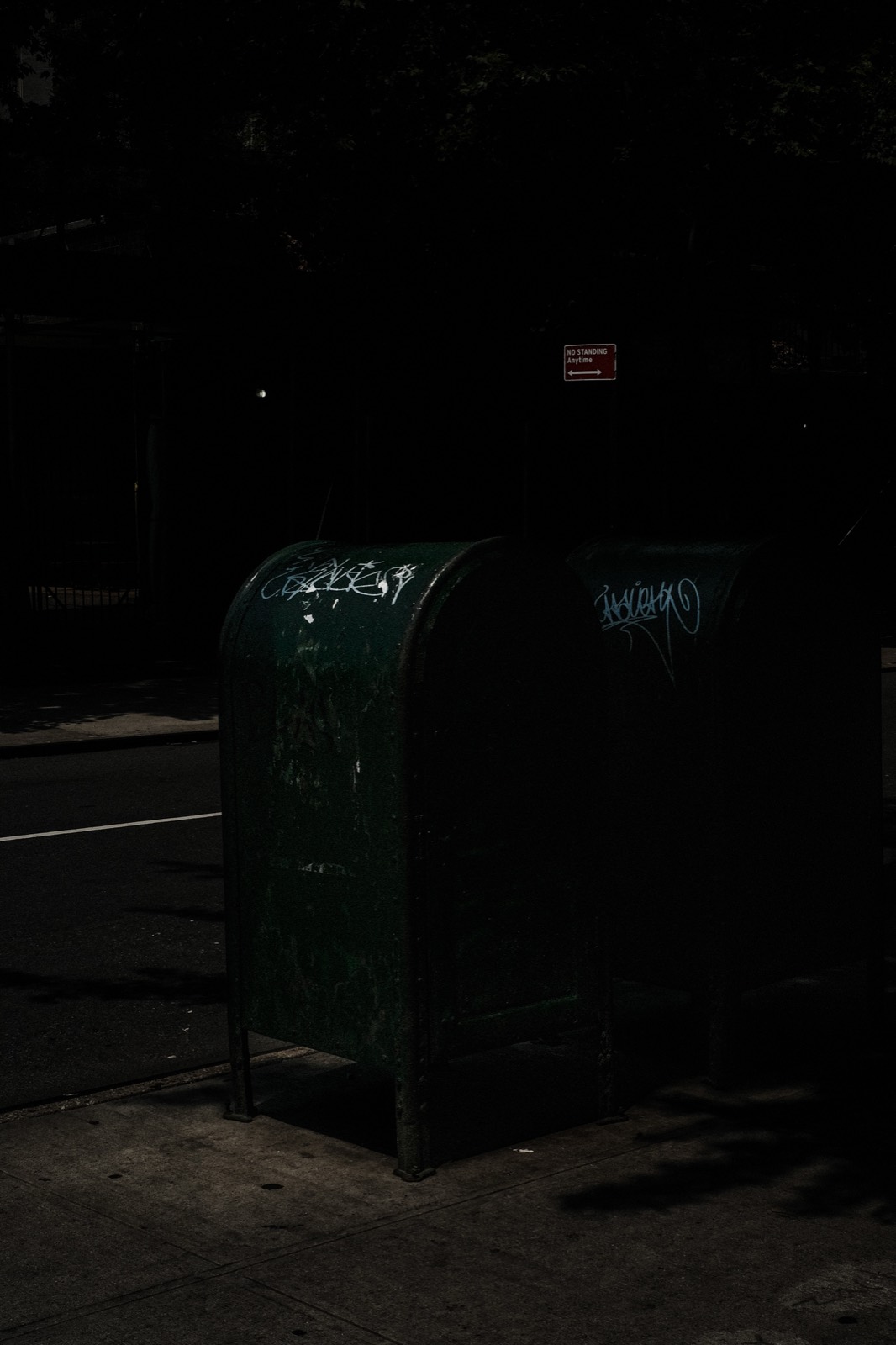 Two green postal relay boxes catching a strip of sunlight on a dark New York sidewalk.
