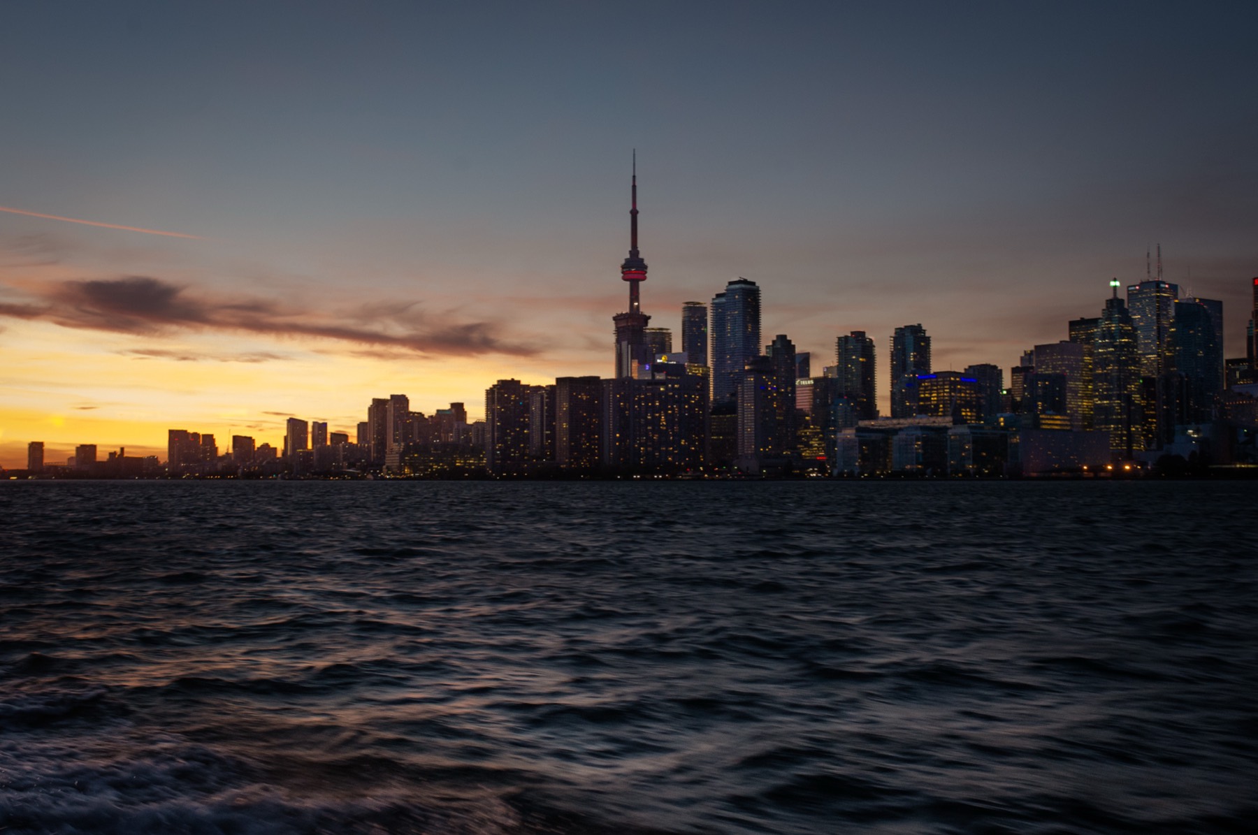 The Toronto skyline and CN Tower at dusk, seen from the harbour with an amber sunset.